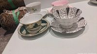 Four fine china tea cups and saucers arranged on a table showing different designs including white with leaf pattern, pink with gold trim, and black and white pattern.