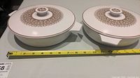 Pair of round white casserole dishes with lids, featuring gold and brown decorative floral patterns on top of the lids, with measuring tape showing approximate 9 inch width.