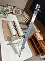 Photo of the aluminum portable artist easel standing on a white table next to a tan fabric tote bag and a rolled white sheet secured with a black elastic band.