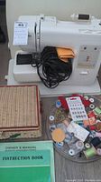 Photo showing Kenmore sewing machine with power cord coiled on top, wicker box, round organizer with assorted sewing accessories, and green instruction manual.
