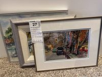 Two framed paintings leaning against a wall on carpet, one watercolor painting by Neal Navarro in front, second larger muted tone oil painting behind
