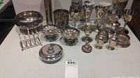Collection of silver plated tableware on white surface with various goblets, salt and pepper shakers, toast rack, small bowls, and covered dish.