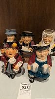 Photo showing five Toby jugs of various characters arranged close together on a wooden surface for lot overview.