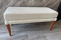 Side angle view of the bench showing the beige patterned upholstery and wooden curved legs.