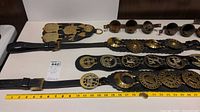 Three black leather straps displayed with attached brass bells and decorative brass medallions, laid horizontally on a white table with yellow measuring tape nearby