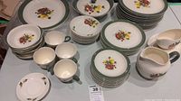 Overview of the dinnerware assortment showing plates, cups, creamer, and gravy boat with fruit design and green rims.