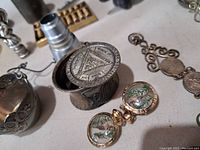 Close-up photo of decorative metal lidded box with geometric patterns, pair of cufflinks with fish paintings, and other metal items in the background