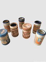 Seven Edison phonograph record cylinders with original cylindrical cardboard containers in various colors and wear.