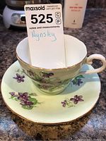 Tea cup and saucer showing violet flowers and gold trim on a light background.