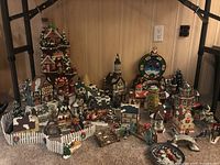 Wide view of clustered Christmas village houses, figurines, fences, faux trees, and mantel clock on a carpeted floor against a wood-paneled wall