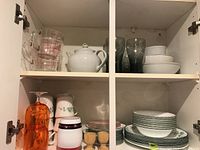 Open cabinet shelf with dishes on bottom and drinkware on top. Bottom shelf contains Corelle dishes with green stripes, bowls, Coca Cola glasses, orange acrylic goblets and wooden egg cups. Top shelf contains Pyrex measuring cups and white ceramic teapot.