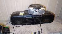 Two audio players placed on carpet floor near wall: a larger black Panasonic cassette/CD player and a smaller silver Sylvaniania CD player on top.