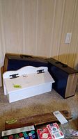 Wood mailbox painted white with black hinges and an accordion file folder placed on carpet against a wall with wood paneling.