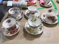 Five Royal Albert tea cups and saucers grouped on a table covered with brown paper. Various floral patterns and gold trim on each piece visible.
