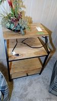 Front angled view of the wood and metal table showing the drawer, bottom and middle shelves, and outlet on the top shelf with a power cord.