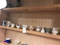 Shelf showing eleven assorted tea cups and mugs arranged side by side, varying in patterns and styles from floral to gold trimmed.