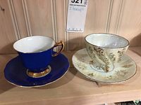 Two tea cups and saucers, one cobalt blue Gladstone and one Japanese floral, displayed on wooden shelf.