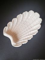 Top and angled view of white ceramic shell-shaped dish showing detailed textured ridges, curled edge, and glossy finish.