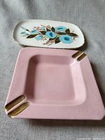 Two trays on fabric background, pink ashtray showing corner gold accents, floral tray with blue flowers and signed mark