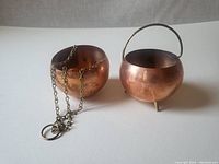 Two small copper planters side by side on a white surface, one with a chain for hanging and one with legs and a handle.