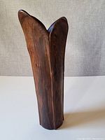 Front view of tall handcarved wooden vase on white surface with light gray fabric background, showing its curved petal-like opening and natural wood grain.