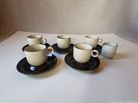 Full set of five ceramic cups on matching dark brown saucers, and small milk pitcher, displayed on white surface against neutral background.