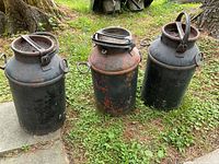 Three vintage metal milk cans arranged side by side on grass with concrete slab on side, showing overall condition and paint