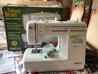 Sewing machine front and side view with original box and manual behind it, presser foot and bobbins in plastic bags placed on machine base.