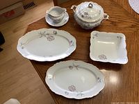 Full set of five fine china serving pieces arranged on wooden table, showing two oval platters, square bowl, tureen with lid, and gravy boat.