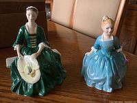 Two Royal Doulton porcelain figurines displayed on a wooden table, showing the Lady in green dress and the Child in blue dress side by side.