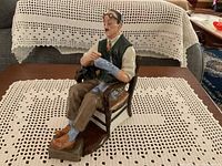Front and side view showing the male figure seated in a wooden chair darning his sock, highlighting detailed paint and textures.