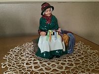 Front view of the Royal Doulton figurine dressed in green checkered shawl, holding colored ribbons, seated on bench with silks beside her.