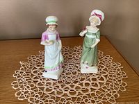 Front view of two Royal Doulton Kate Greenway series figurines: Ruth in green dress and Beth in mauve dress, displayed on a lace doily.