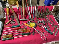 Overview of mixed hand tools including hammers, pliers, screwdrivers, files, sockets, wrenches, measuring tape, rulers, and small miscellaneous tools laid out on a red cloth.