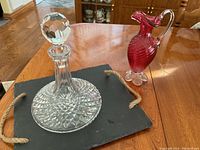 Waterford crystal captain decanter with stopper, cranberry swirl glass pitcher with clear handle and base, and square slate serving tile with rope handles arranged on a wooden table.
