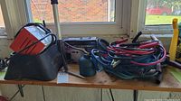 Photo showing Motomaster car battery charger, grease tools, and coiled jumper cables on a wooden table near a window
