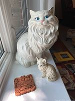 Two white ceramic cats and one small terracotta plaque on a white surface near window light.