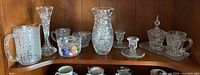 Wide view of all crystal items including pitcher, candlesticks, creamer, sugar bowl, various small bowls and vases arranged on wooden shelf.