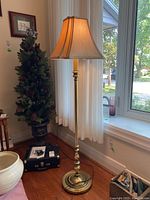 Full height image of the floor lamp showing its brass finish and beige fabric shade near a window and hardwood floor.