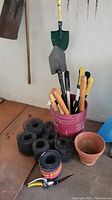 Red bucket filled with garden tools such as shears and wooden handles, five rolls of black garden edging, two terra cotta flower pots
