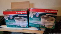 Two Honeywell humidifiers in their original bright red and white boxes placed side by side on a wooden shelf in a basement setting.