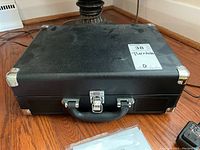 Closed black portable turntable case with metal reinforced corners and a single latch in the center with carrying handle on wooden floor.