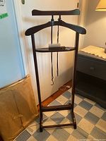 Full front view of the wooden clothing valet showing hanger, shelf with brush and shoehorn, cords hanging from the shelf.