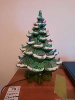 Front view of vintage green ceramic Christmas tree with white snow tips and colorful bulbs on branches placed on wooden surface with price ticket showing 'Ceramic Christmas Tree'.