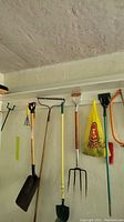 Photo showing variety of yard tools including shovels, a garden fork, hard rake, some smaller tools, and a bow saw, all hanging on hooks on a wall.