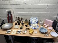Full view of all items on table showing assortment of decorative and collectible pieces including bowls, figurines, clock plate, and pink boxes.