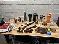 Photo of assorted household and personal items on wood table including drinkware, sunglasses, and decorative pieces