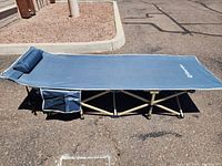 Blue camping cot fully assembled outdoors on asphalt ground, showing side profile with integrated pillow and side pocket under the head area.