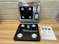 Photo showing the Fine Life smart weight scale in front of its original box and an instruction sheet with smartphone QR codes.