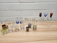 Wide view of the lot showing an assortment of shot glasses and mini wine glasses arranged side by side on a wooden surface against a white brick wall.